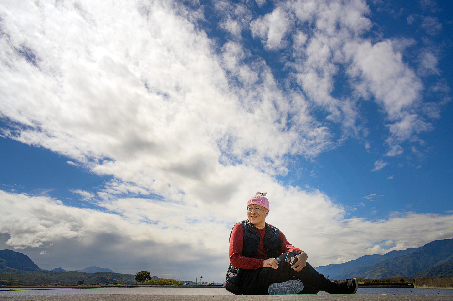 Dzongsar Khyentse Rinpoche sits on the ground wearing a pink hat with a big blue sky behind him.